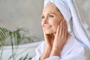 Older female waring a robe and towel on her head. Touching her cheek with her hands as if apply a skincare product>